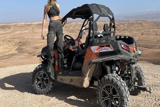 Two people driving a 1000cc buggy through the Agafay Desert with rocky trails and the Atlas Mountains in the background